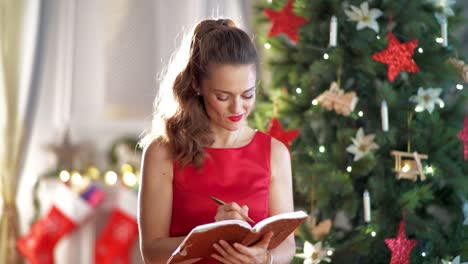 ama-de-casa-sonriendo-cerca-de-árbol-de-Navidad-de-la-escritura-en-el-cuaderno