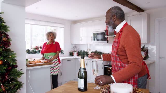 Senior-schwarzer-Mann-gießt-Champagner-für-seine-Partnerin-in-der-Küche,-während-sie-am-Weihnachtstag-Abendessen-vorbereiten