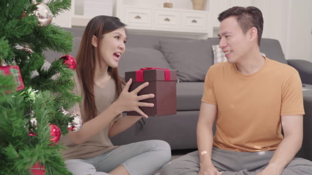 Asian-couple-man-giving-christmas-gifts-to-his-girlfriend-in-their-living-room-at-home-in-Christmas-Festival.-Lifestyle-woman-and-man-happy-celebrate-summer-christmas-and-New-year-concept.