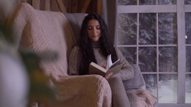 Young-girl-with-long-hair-reads-a-book-while-sitting-in-a-chair-near-the-Christmas-tree.