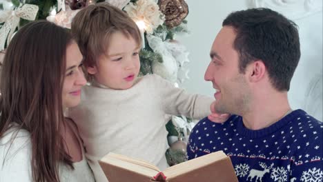 Happy-father-reading-about-Santa-beard,-while-mother-holding-their-playful-son-in-front-of-the-Christmas-tree