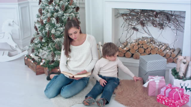 Junge-Mutter-liest-Buch,-um-ihren-Sohn-von-Weihnachtsgeschenken-abzulenken