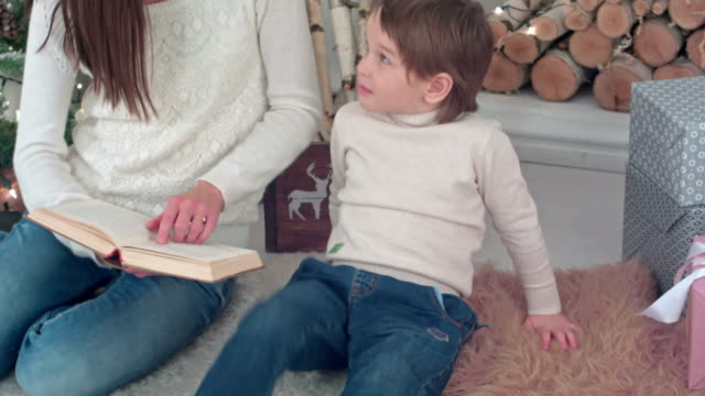 Chico-caprichoso-escuchando-a-su-mamá-leyendo-un-libro-cerca-de-árbol-de-Navidad