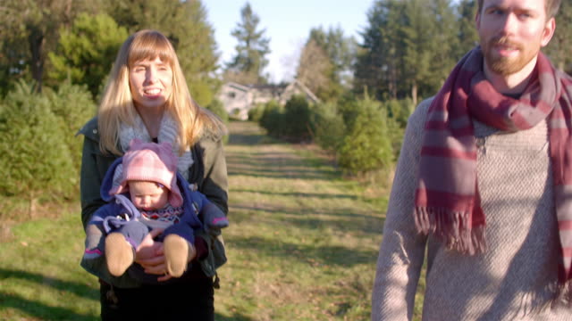 Una-joven-familia-caminando-a-través-de-una-granja-de-árboles-de-Navidad,-madre-que-lleva-bebé