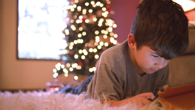 Little-boy-laying-on-the-floor-using-his-tablet,-Christmas-tree-with-lights-behind-him