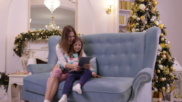 Mother-With-Daughter-On-Couch-Using-Tablet-Computer-Happy-Smiling-Young-Family-Near-Decorated-New-Year-Christmas-Tree