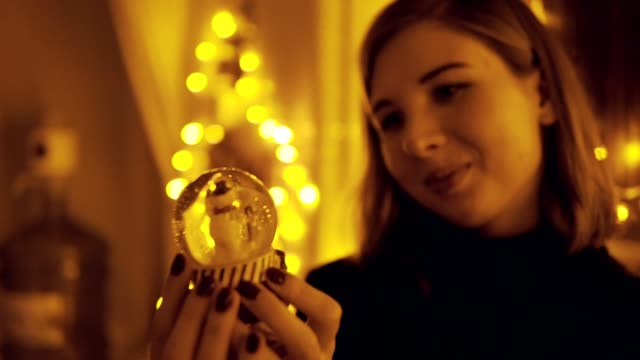 Portrait-of-a-young-woman-with-fairy-lights-in-the-house-shaking-the-snow-globe