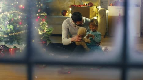 Blick-durch-Schneefenster-Sitzen-unter-Weihnachten-Drei-Vater-und-Tochter-spielen-mit-Plush-Bär