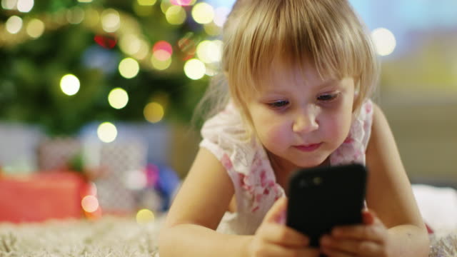 Niedliche-kleine-Mädchen-mit-Smartphone-in-der-Hand-liegt-auf-dem-Teppich-unter-dem-Weihnachtsbaum.