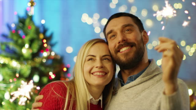 Glückliches-Paar-leichte-Wunderkerzen-und-Lächeln.-Im-Hintergrund-Weihnachtsbaum-und-Zimmer-mit-Lichtern-geschmückt.