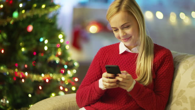 Hermosa-mujer-rubia-se-sienta-en-un-sofá-y-aplicaciones-Smartphone.-Árbol-de-Navidad-y-decoradas-con-luces-son-en-el-fondo.