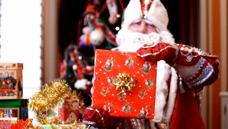 Canta-Klaus,-Father-Christmas,-Father-Frost-hält-in-seinen-Händen,-in-Fäustlinge-ein-großes-Weihnachtsgeschenk-in-einer-roten-Papierverpackung-mit-einer-goldenen-Schleife,-zeigt-mit-seiner-Hand-ok,-im-Hintergrund-ist-ein-Weihnachtsbaum