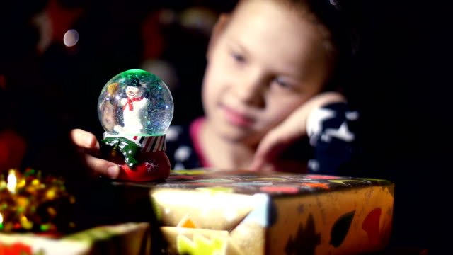 In-the-twilight-of-the-night,-among-the-gifts-in-bright-colorful-paper-packages,-a-pretty-little-blonde-girl-with-a-pink-bow-in-her-hair-and-a-beautiful-dress,-looks-carefully-and-admires-snow-globe.-toy-Christmas-ball-with-sparkles-and-a-snowman-inside