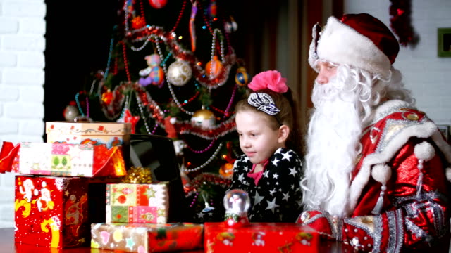 In-the-twilight-of-the-night,-among-the-gifts-in-bright-colorful-paper-packages,-the-pretty-blonde-girl-with-a-pink-bow-in-her-hair-and-in-a-beautiful-dress-and-Santa-Claus,-Father-Christmas,-Father-Frost-watching-something-cheerful-on-a-laptop.-They-talk