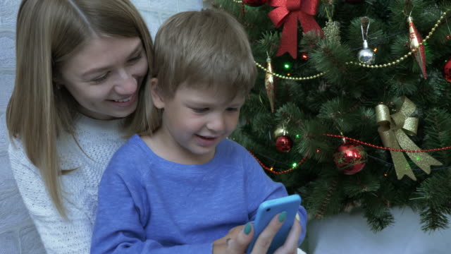 Fröhlicher-Junge-und-seine-Mutter,-Blick-auf-das-Smartphone-in-der-Nähe-von-Christmas-tree