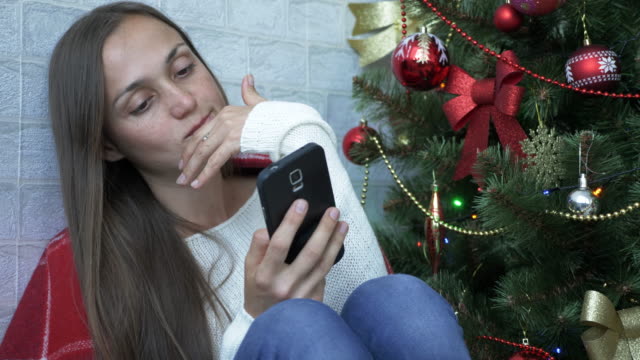 Frau-sitzt-auf-einer-Etage-mit-roten-Plaid-und-mit-Smartphone-neben-Weihnachtsbaum