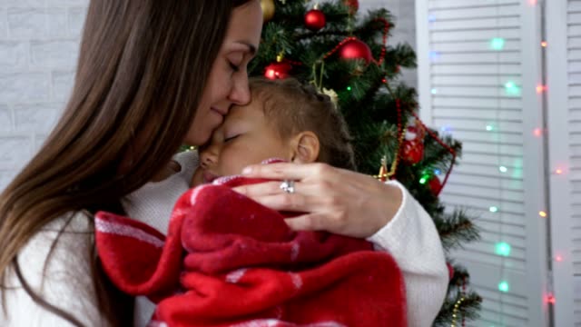 Young-mother-holding-her-sleeping-little-daughter-in-hands-near-Christmas-tree.