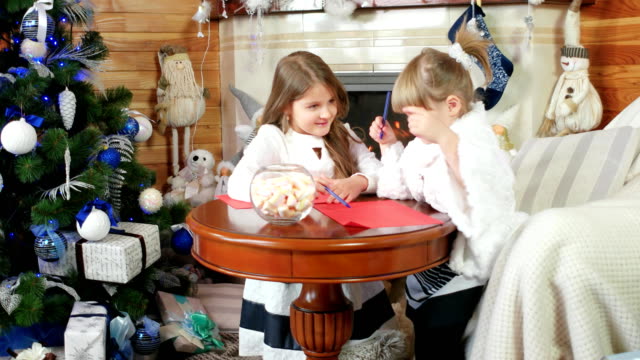 Letter-for-Santa-Claus,-two-girls,-sisters-write-and-draw,-girls-sit-near-the-fireplace,-children-in-the-house-near-Christmas-tree