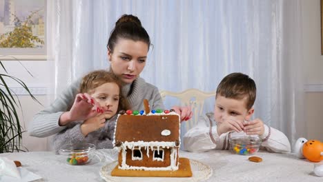 Junge-Mutter-mit-zwei-Kindern-schmückt-ein-Lebkuchenhaus-mit-einem-Bonbons