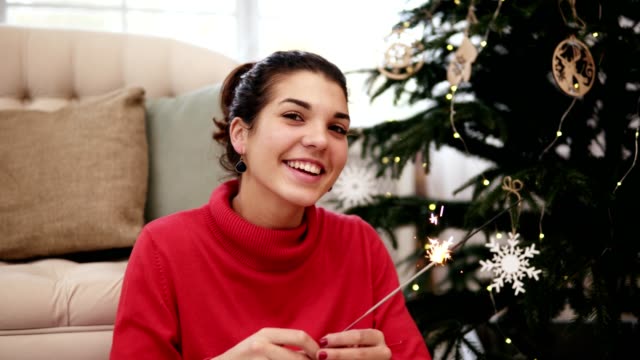 Freundliche-attraktive-junge-Frau-sitzt-auf-dem-Boden-durch-den-Weihnachtsbaum-feiert-Weihnachten-mit-Wunderkerzen-und-schaut-in-die-Kamera-Lächeln