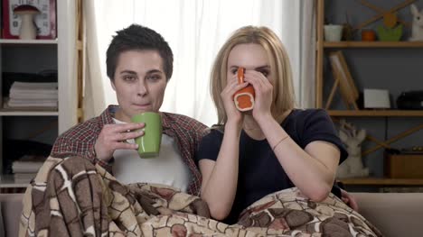 Two-young-lesbian-girls-are-sitting-on-the-couch,-covered-with-a-warm-blanket,-holding-cups-in-their-hands,-drinking-dark-tea,-coffee,-cuddling,-looking-at-the-camera-60-fps