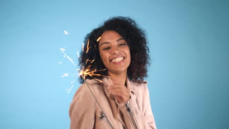 Sparkling-Bengal-fire-in-african-american-woman's-hand-on-blue-background.-Christmas-Holiday-Concept.-Young-pretty-girl-with-afro-hairstyle-celebrating,-smiling,-enjoying-time.-4k