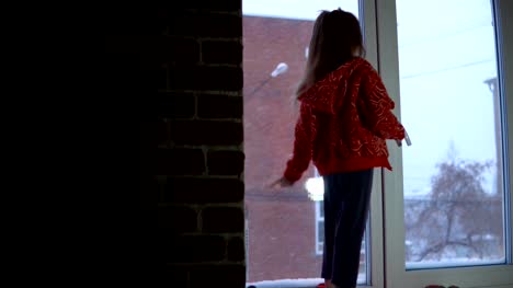 Little-cute-girl-standing-on-the-window-sill,-looking-out-on-a-snowy-cityscape.