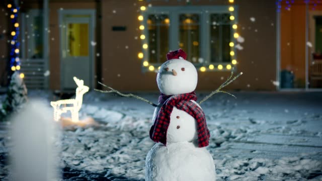 Funny-Snowman-Wearing-Hat-and-Scarf-Standing-in-the-Backyard-of-the-Idyllic-House-Decorated-with-Garlands-on-Christmas-Eve.-Soft-Snow-is-Falling-on-that-Magical-Winter-Evening.