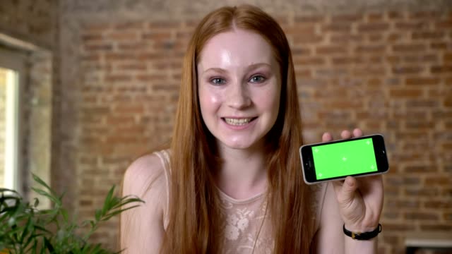 Young-fresh-ginger-girl-is-showing-green-screen-on-her-smartphone,-watching-at-camera,-communication-concept,-brick-background