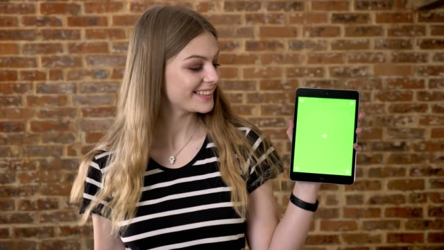 Joven-blonda-feliz-muestra-pantalla-verde-de-la-tableta,-como-signo,-concepto-de-comunicación,-el-fondo-de-ladrillo