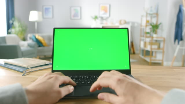 First-Person-Close-up-Shot-Man-Uses-Laptop-with-Green-Mock-up-Screen-While-Sitting-at-the-Desk-in-His-Cozy-Living-Room.