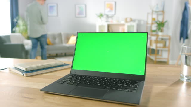 Modernen-Laptop-mit-grünen-Mock-up-Display-auf-dem-Schreibtisch-in-das-gemütliche-Wohnzimmer-stehen.-Mann-mit-Handy-sitzt-auf-einem-Stuhl,-Rest.