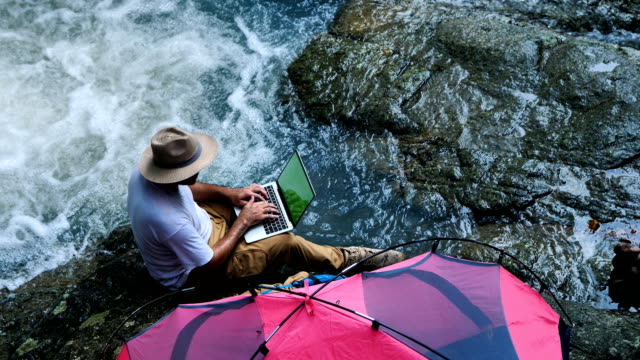 Joven-usando-la-computadora-portátil-de-pantalla-verde-en-una-cascada-y-un-Camping.-Concepto-de-trabajo-viaje-y-Freelance