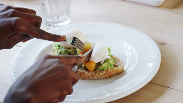 Nahaufnahme-der-Hände-mit-Messer-und-Gabel-in-ein-weich-gekochtes-Ei-mit-Avocado-auf-Toast,-serviert-auf-einem-weißen-Teller-im-Restaurant,-Detail,-selektiven-Fokus-schneiden