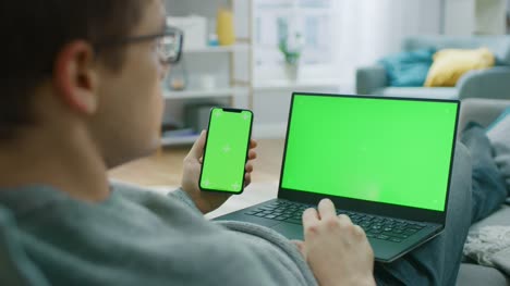 Young-Man-at-Home-Works-on-a-Laptop-Computer-with-Green-Mock-up-Screen,-while-Holding-Smartphone-with-Chroma-Key-Display.-He's-Sitting-On-a-Couch-in-His-Cozy-Living-Room.-Over-the-Shoulder-Camera-Shot