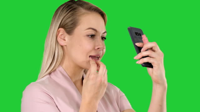 Beautiful-young-blonde-woman-which-is-painting-her-lips-with-red-lipstick-and-looking-in-the-phone-on-a-Green-Screen,-Chroma-Key