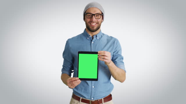 Mid-Shot-of-a-Fashionable-Young-Man-in-a-Hat-and-Glasses-Presenting-to-Us-Tablet-Computer-with-Isolated-Green-Screen.-Schuss-auf-einen-weißen-Hintergrund.