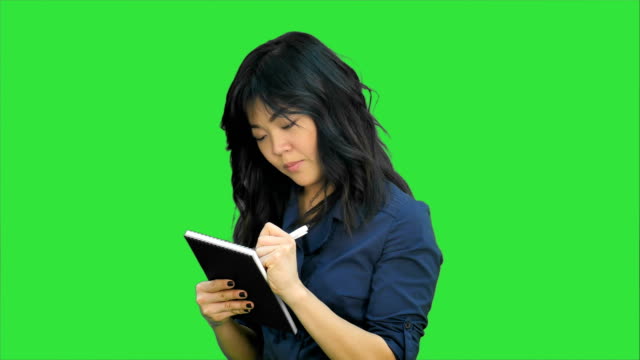 Pensive-asian-woman-makes-some-notes-in-notepad-on-a-Green-Screen,-Chroma-Key