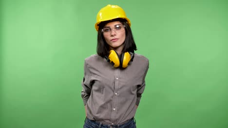 Young-pretty-builder-woman-standing-in-earphones,-glasses-and-helmet-with-hands-in-pockets-and-looking-in-camera,-chroma-key-background