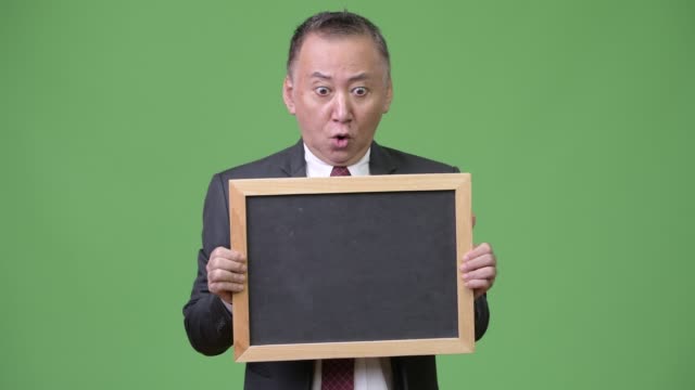 Reife-japanischer-Geschäftsmann-hält-Tafel-vor-grünem-Hintergrund