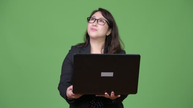 Mature-beautiful-Asian-businesswoman-using-laptop-against-green-background