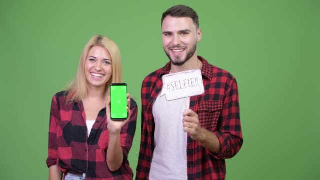Young-couple-showing-phone-while-holding-selfie-paper-sign-together