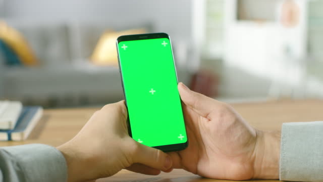 Close-up-of-a-Man-Holding-Green-Mock-up-Screen-Smartphone-in-His-Hands.-Using-Wireless-Device-to-Browse-Through-Internet.-In-the-Background-Cozy-Living-Room-or-Home-Office.