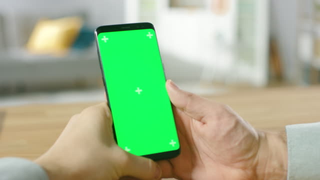 Close-up-of-a-Man-Holding-Green-Mock-up-Screen-Smartphone-in-His-Hands.-Using-Wireless-Device-to-Browse-Through-Internet.-In-the-Background-Cozy-Living-Room-or-Home-Office.