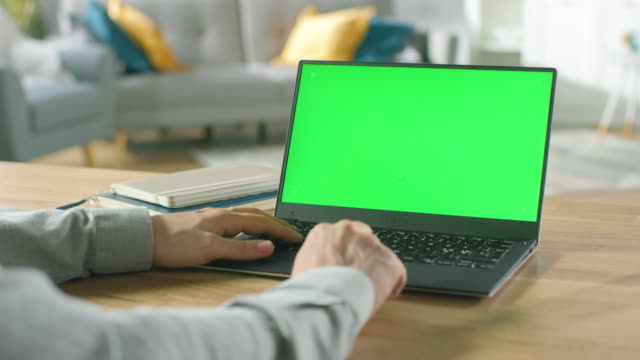 Nahaufnahme-eines-Mannes-verwendet-Laptop-mit-grünen-Mock-up-Bildschirm-sitzend-am-Schreibtisch-in-seinem-gemütlichen-Wohnzimmer.