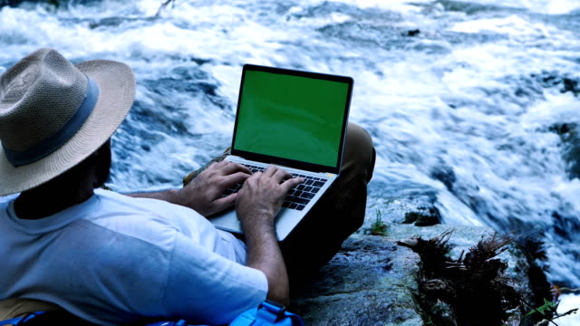 Junger-Mann-mit-green-Screen-Laptop-Computer-auf-einen-Wasserfall.-Reise--und-Freelance-Work-Konzept