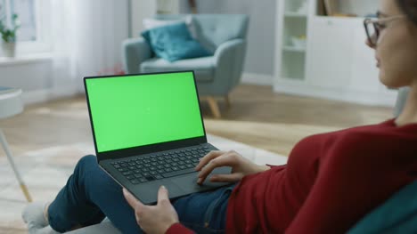 Junge-Frau-zu-Hause-arbeitet-an-einem-Laptop-Computer-mit-grünen-Mock-up-Bildschirm.-Sie-sitzt-auf-einer-Couch-in-seinem-gemütlichen-Wohnzimmer.-Über-die-Schulter-Kamera-Aufnahme