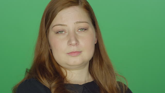 Young-redhead-woman-tries-to-act-serious-but-begins-to-laugh,-on-a-green-screen-studio-background