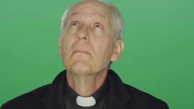 Older-priest-prays-to-the-heavens-and-smiles,-on-a-green-screen-studio-background