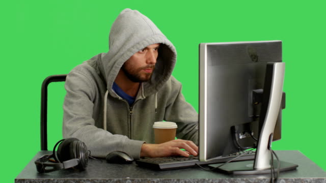 Tiro-medio-de-un-Hacker-con-Hoodie-sentado-en-su-computadora-de-escritorio.-El-fondo-de-pantalla-verde.